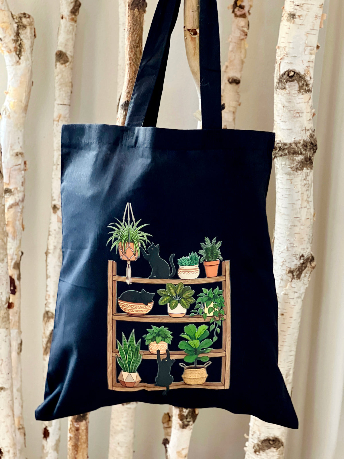Navy tote bag with a graphic of cats and plants on a wooden shelf against a white birch tree background.