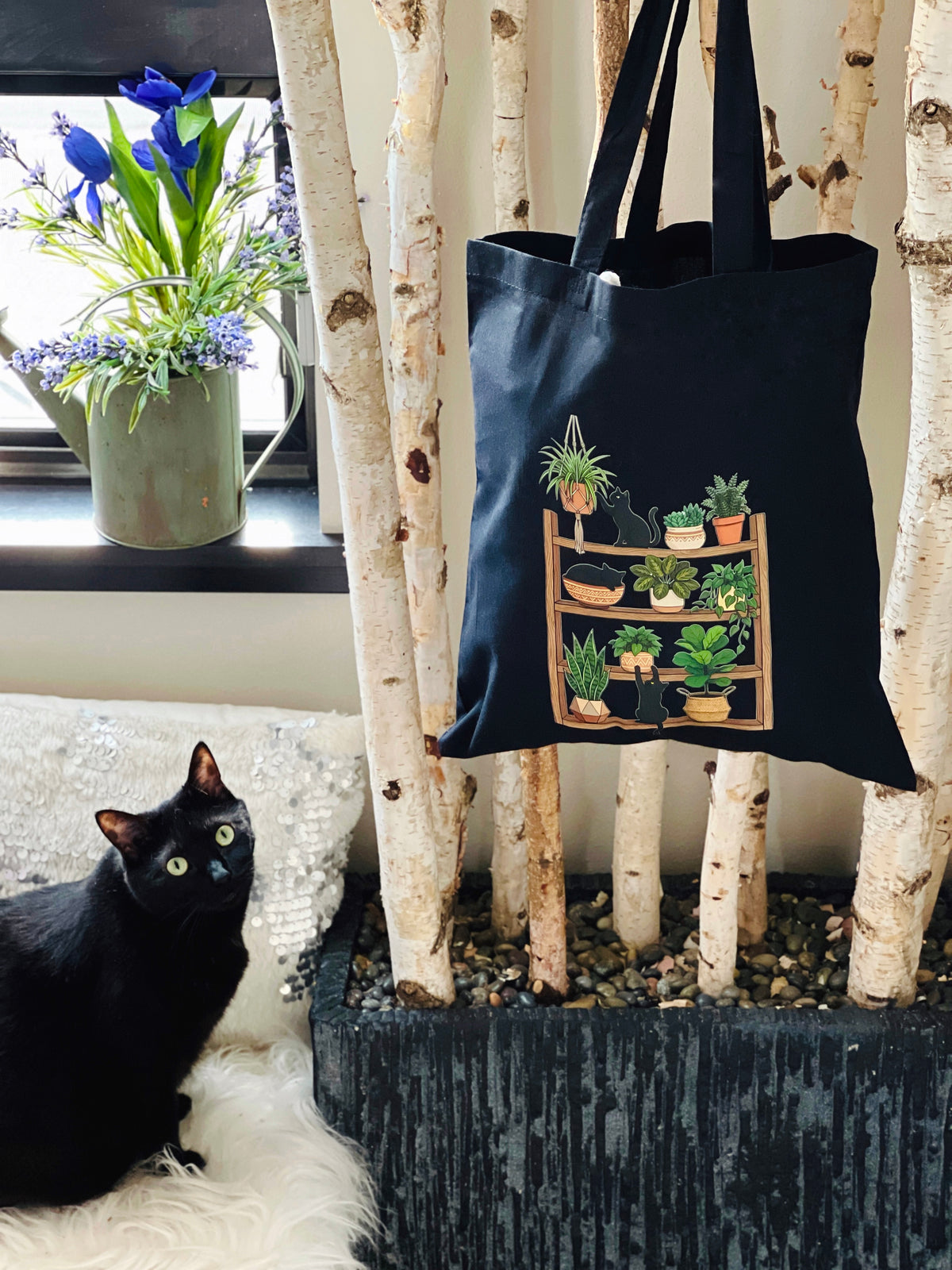 Black tote bag with plant design hanging on a birch branch, black cat sitting on a white surface.