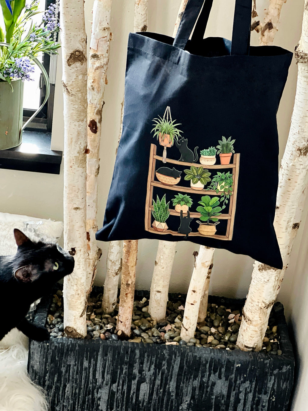 Black tote bag with plant design hanging on a wall next to a black cat.