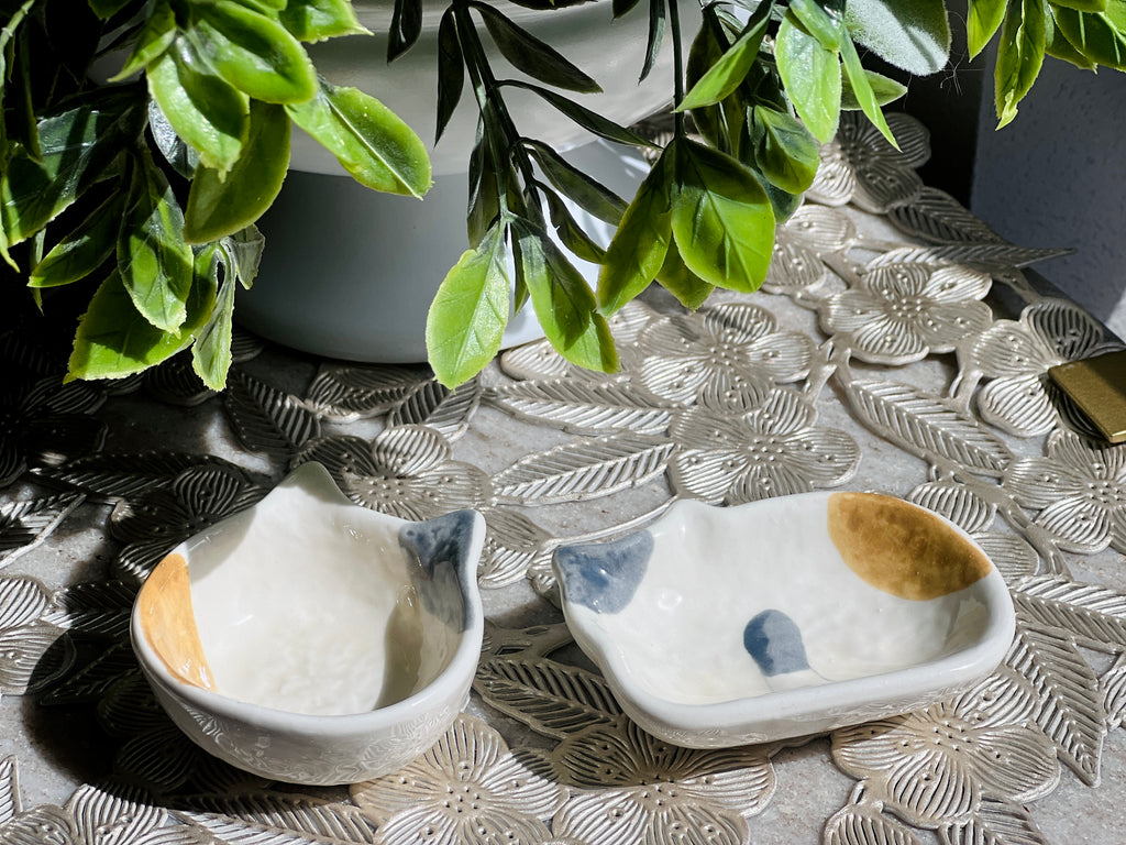 Two small ceramic dishes with cat designs on a decorative surface with plants in the background.