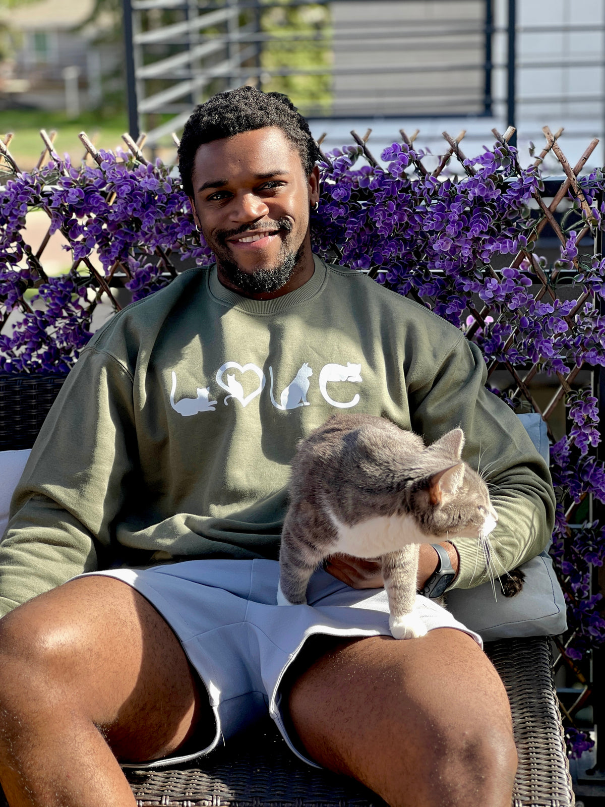Man sitting outdoors with a cat on his lap, surrounded by purple flowers. With "love" cat crewneck.