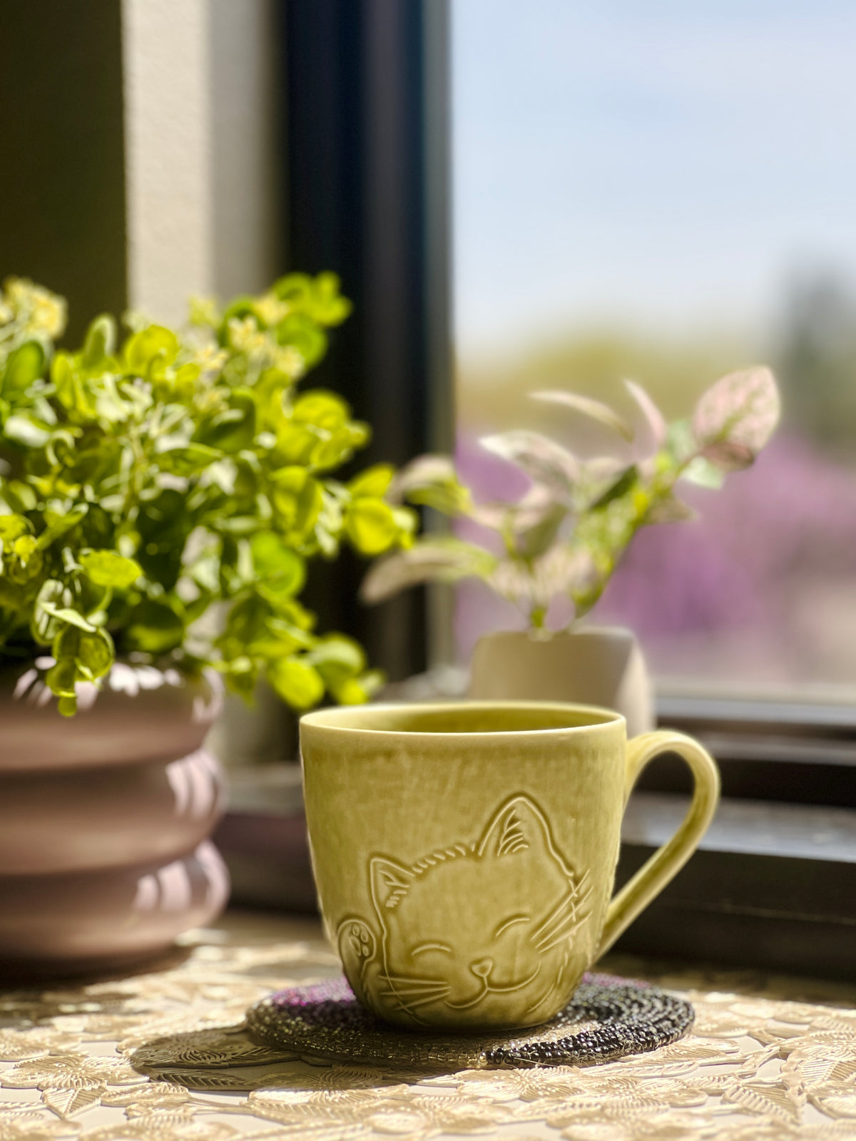 Green ceramic mug with cat design on a table near plants and a window.