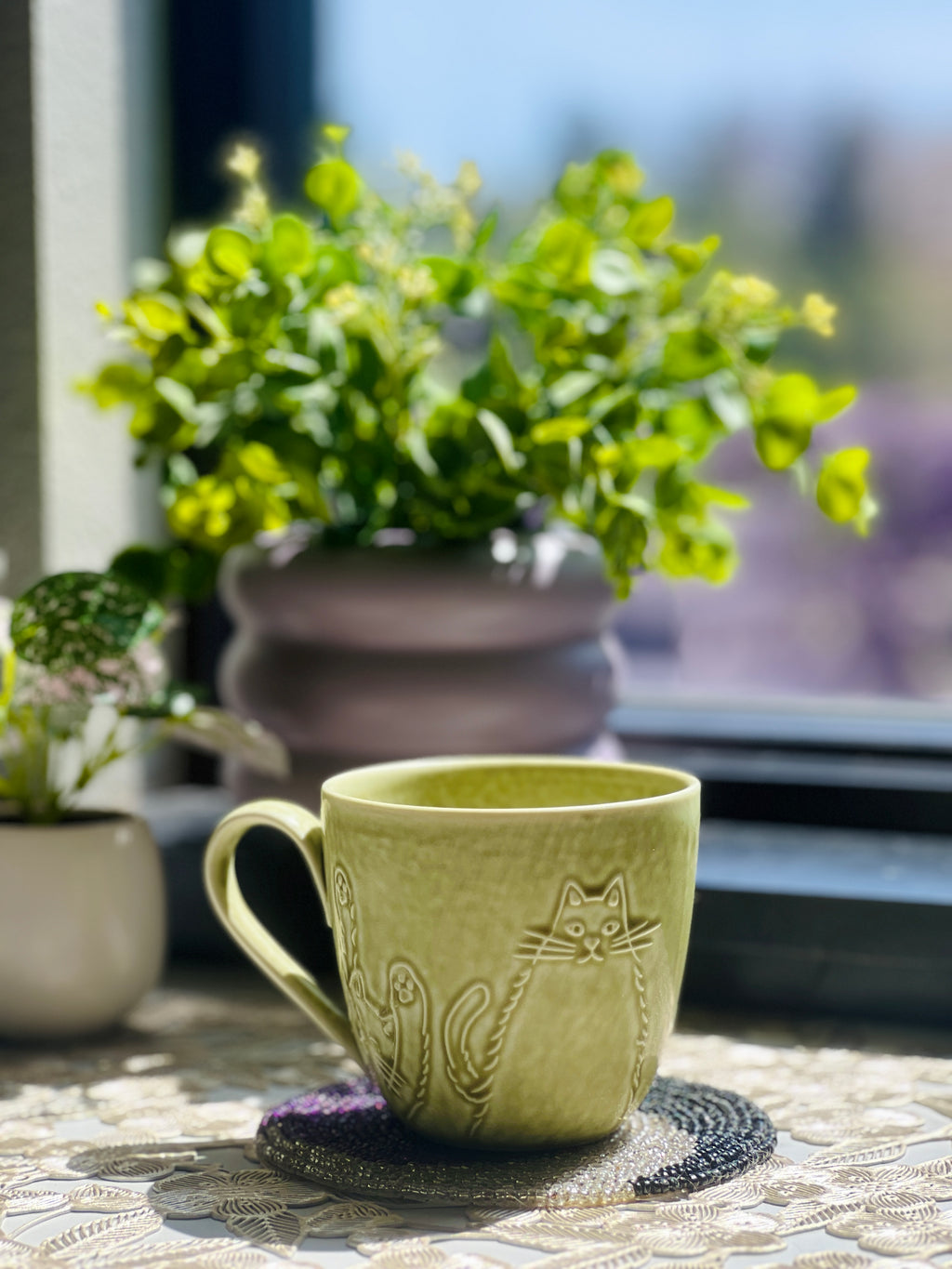 Green ceramic mug with cat design on a table outdoors, surrounded by plants.