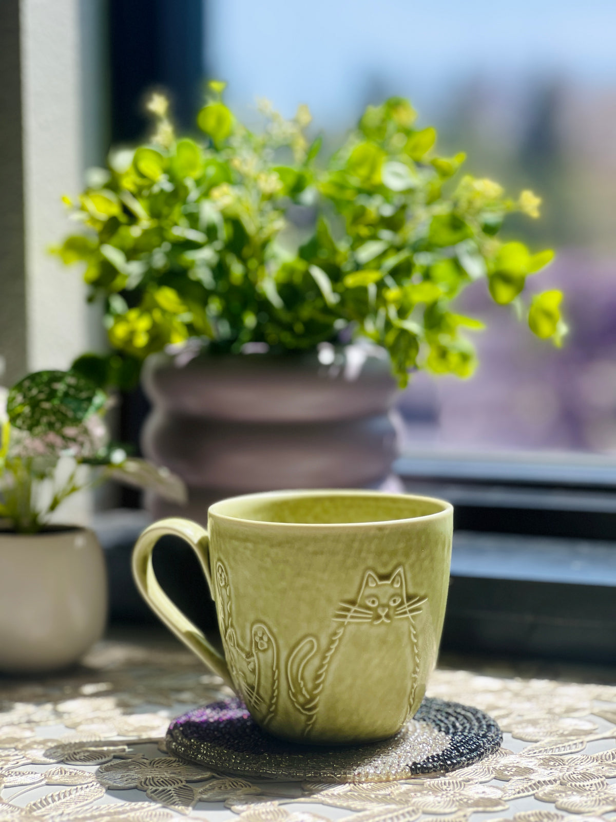 Green ceramic mug with cat design on a table outdoors, surrounded by plants.
