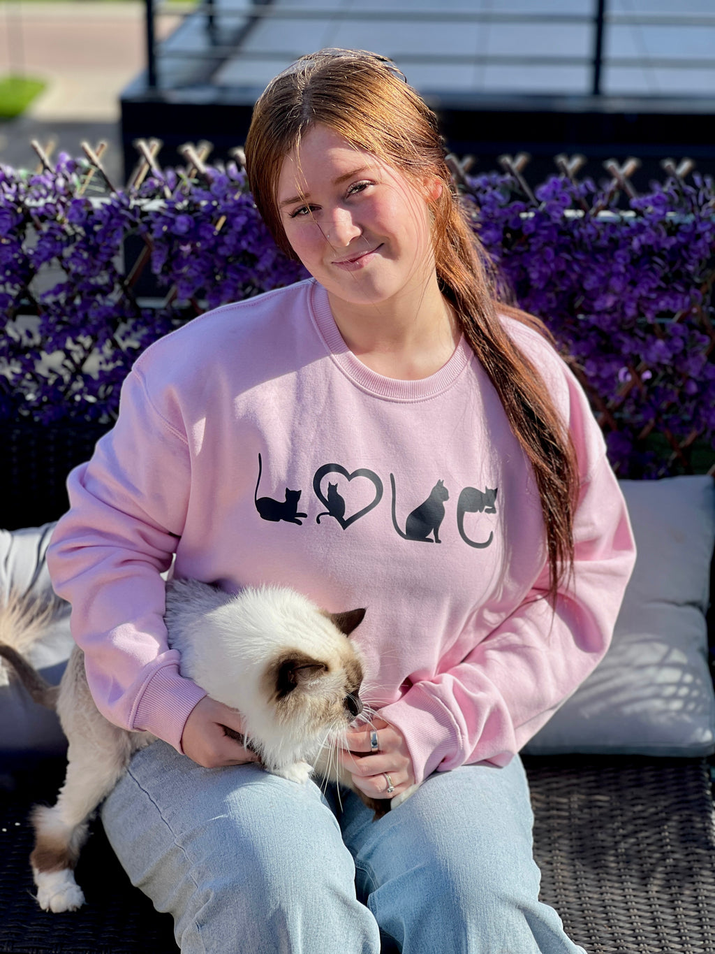 Person wearing a pink sweatshirt with cat designs sitting outdoors with a small white cat.