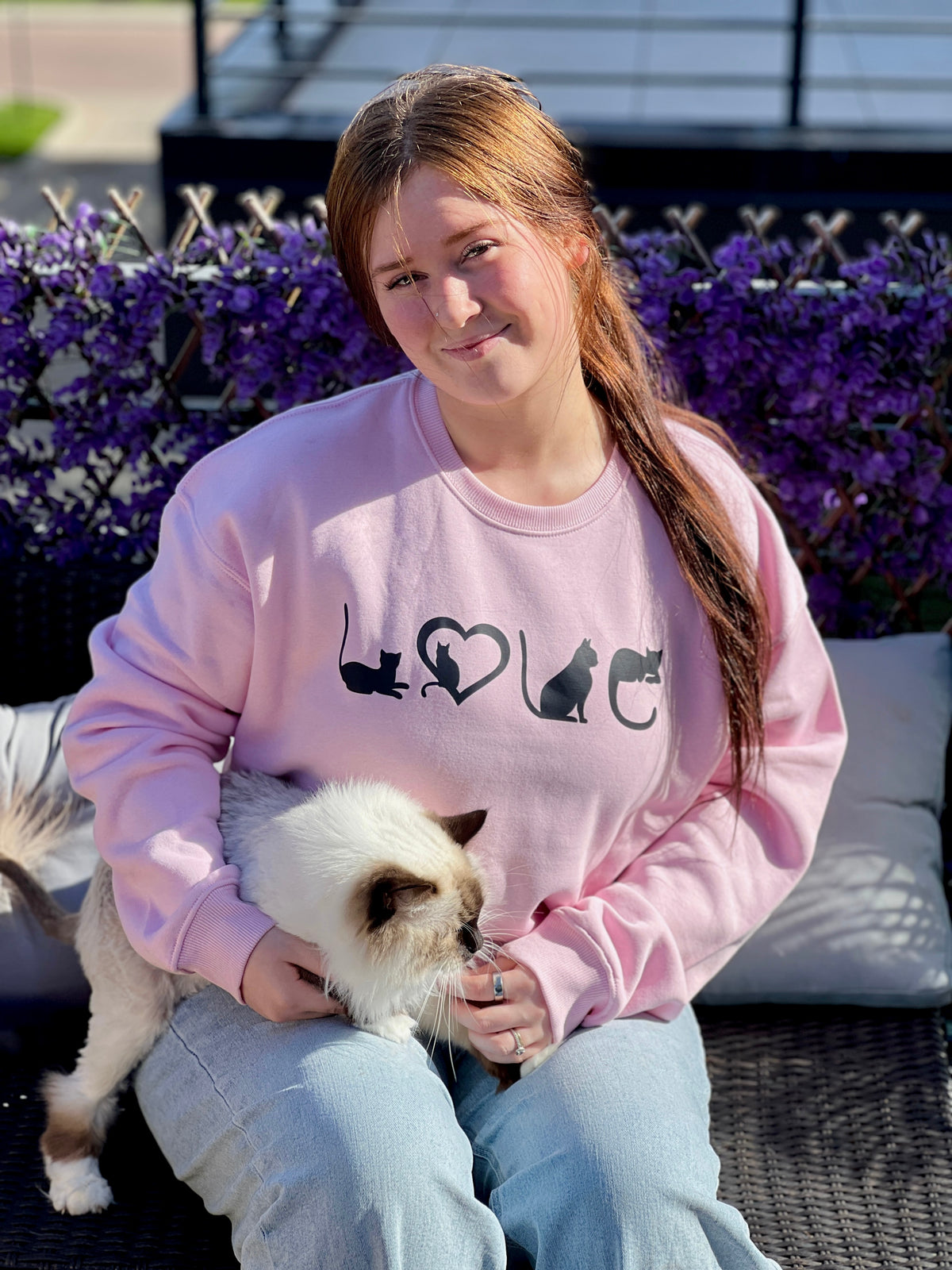 Person wearing a pink sweatshirt with cat designs sitting outdoors with a small white cat.