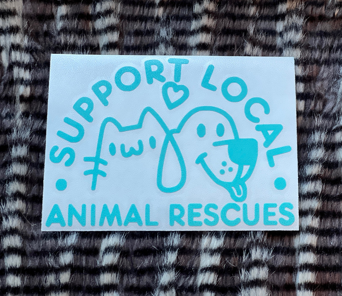 Light blue sticker with text 'Support Local Animal Rescues' featuring a cat and dog illustration on a textured fabric background.