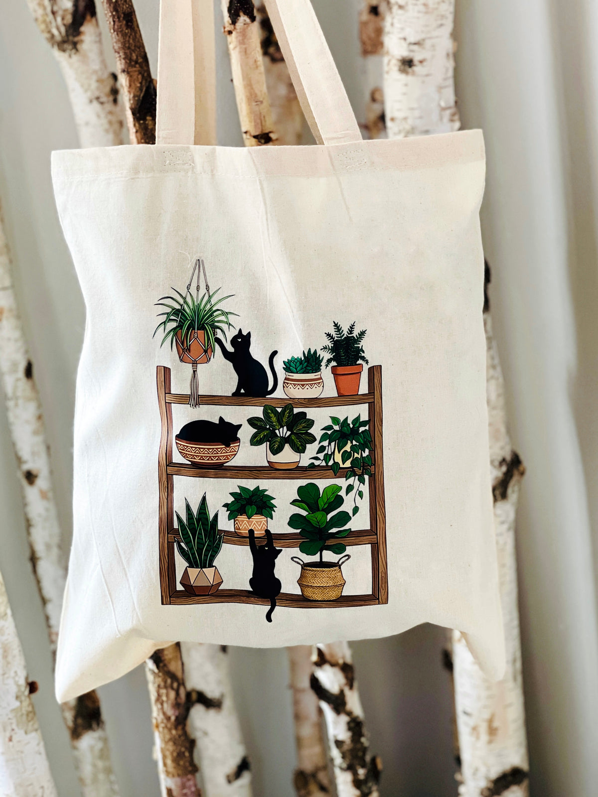 Tote bag with a design of cats and plants on a wooden shelf against a white background