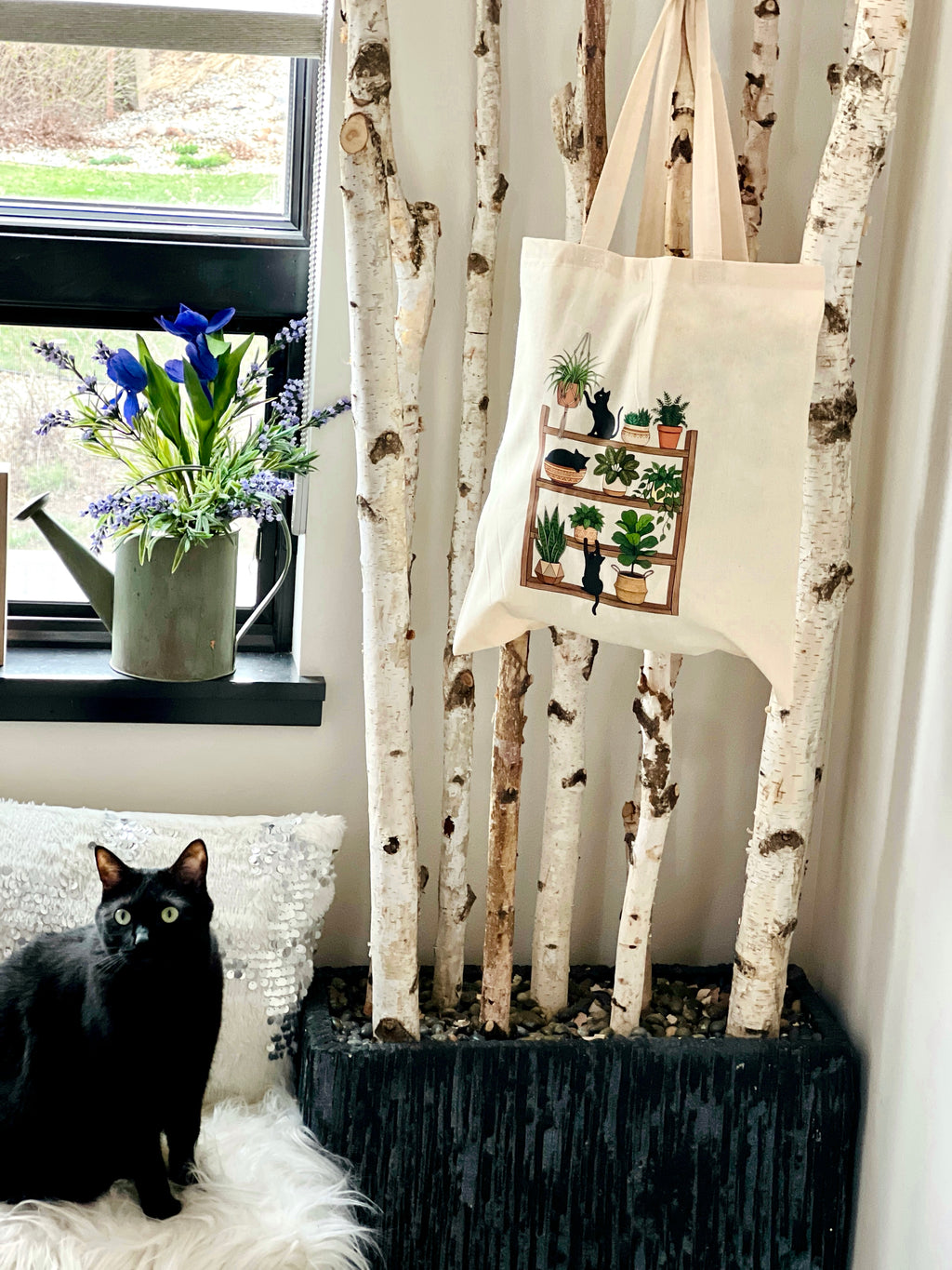 Black cat on a white cushion next to a tote bag with plant designs, in front of birch trees and a window with flowers.