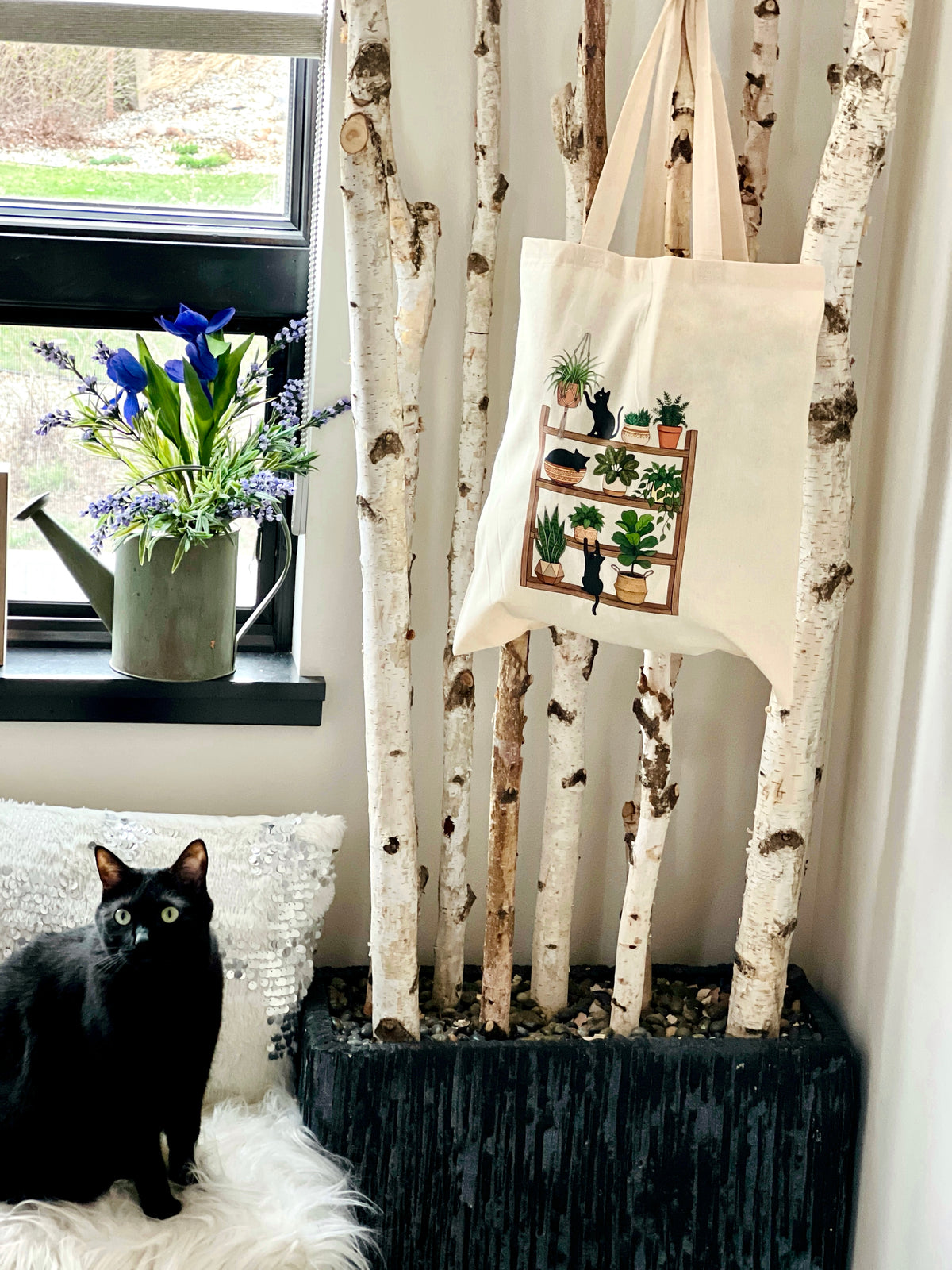 Black cat on a white cushion next to a tote bag with plant designs, in front of birch trees and a window with flowers.