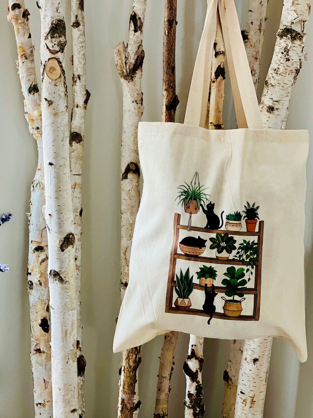 Tote bag with a design of a bookshelf with plants and a cat hanging on birch trees.