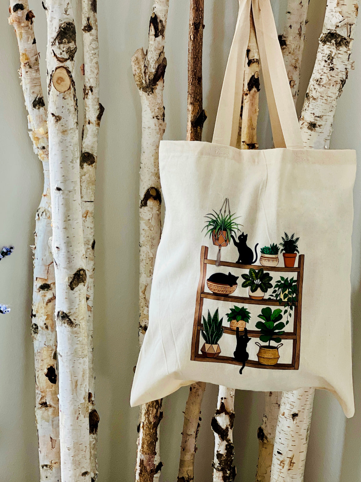 Tote bag with a design of a bookshelf with plants and a cat hanging on birch trees.