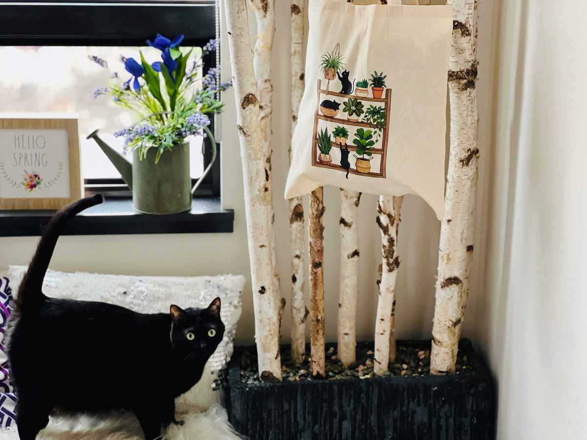 Black cat sitting on a couch next to a tote bag with plant designs, near birch trees and flowers.
